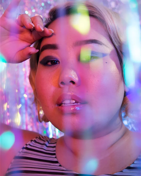 Asian woman poses with graphic eyeliner on rainbow-holographic set.